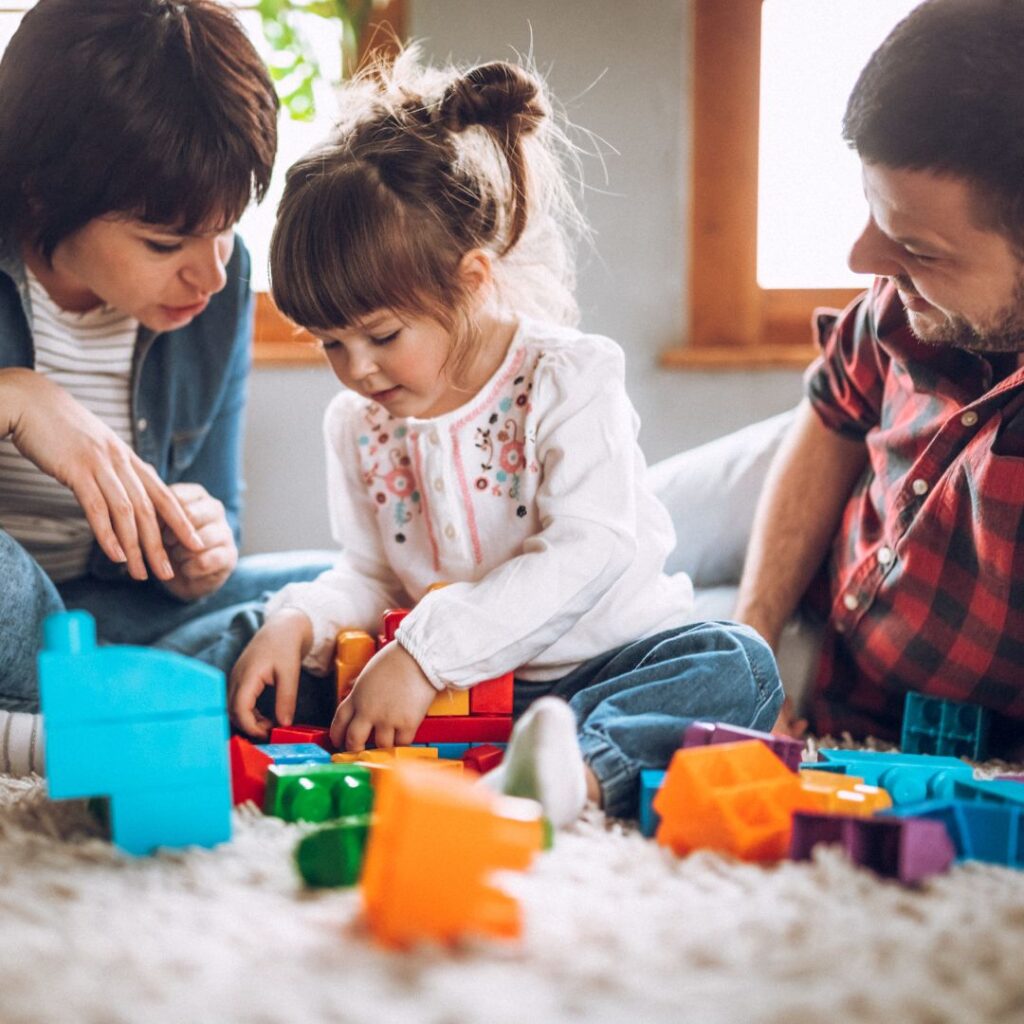 Parents sit on the floor playing Duplo blocks with their preschool aged daughter.